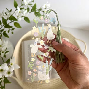 May include: A clear bookmark with a green tassel features a whimsical design of white ducks and pastel flowers. The bookmark is held in a hand against a background of a woven basket and white flowers.