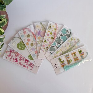 May include: A collection of clear acrylic bookmarks with various designs, including books, flowers, and butterflies. Each bookmark is packaged and features a tassel. The color palette includes pink, green, and blue.
