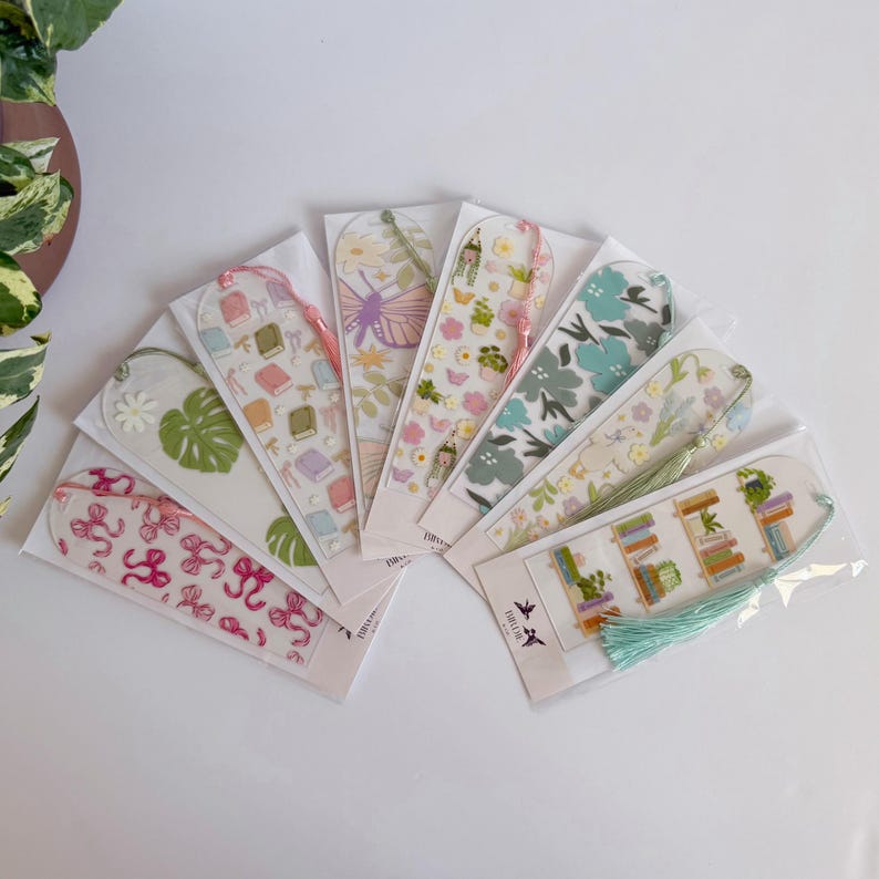 May include: A collection of clear acrylic bookmarks with various designs, including books, flowers, and butterflies. Each bookmark is packaged and features a tassel. The color palette includes pink, green, and blue.
