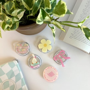 May include: A collection of clear phone grips with various designs, including a yellow daisy, a pink bow, and a goose reading a book. One grip reads "A book a day keeps reality away."