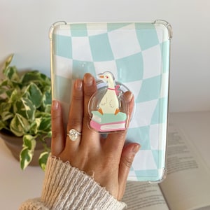May include: Clear phone case with a light blue and white checkered pattern. A clear pop socket with a cartoon goose reading a book is attached. The case is held in front of a plant and an open book.