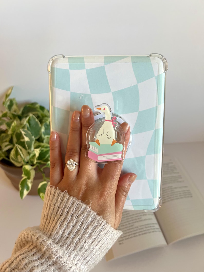 May include: Clear phone case with a light blue and white checkered pattern. A clear pop socket with a cartoon goose reading a book is attached. The case is held in front of a plant and an open book.