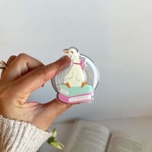 May include: A clear acrylic pin featuring a cartoon goose wearing a pink bow, perched atop two books. The goose has orange feet and a yellow beak. The pin is held in a hand, with a book visible in the background.