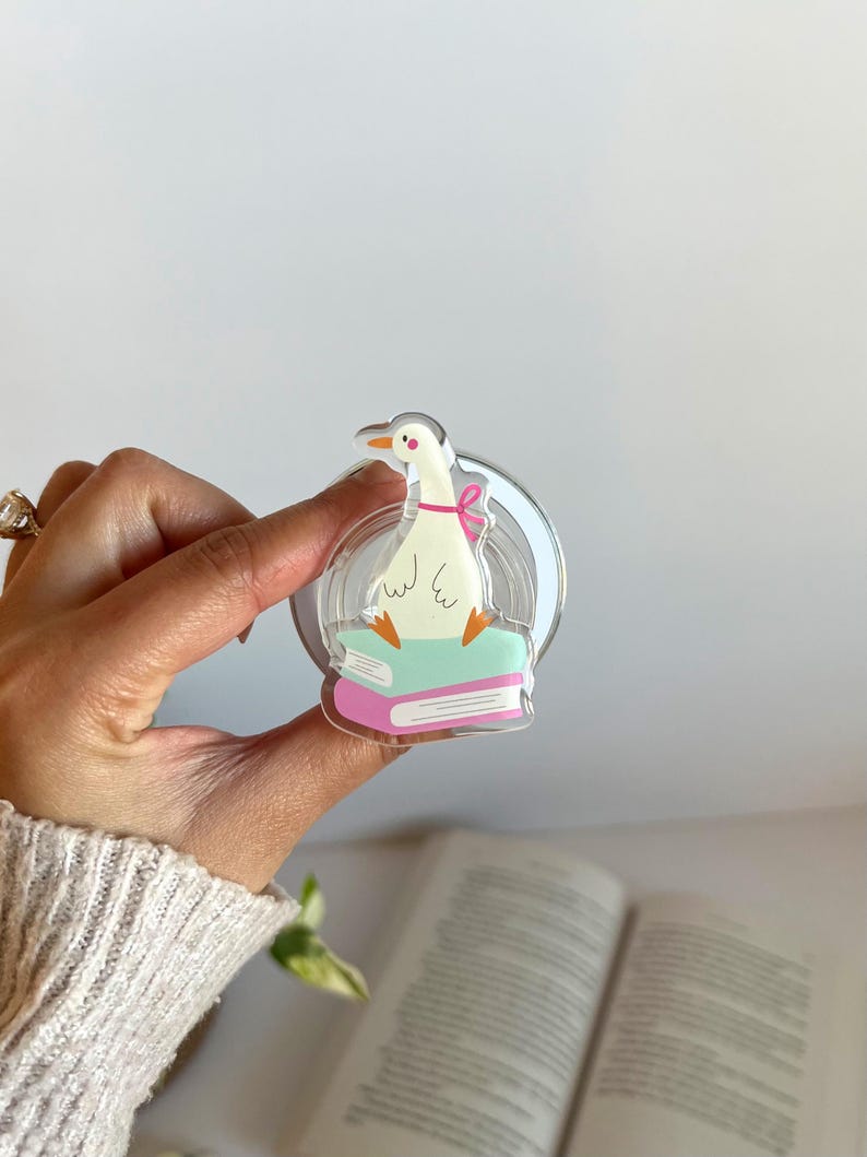 May include: A clear acrylic pin featuring a cartoon goose wearing a pink bow, perched atop two books. The goose has orange feet and a yellow beak. The pin is held in a hand, with a book visible in the background.