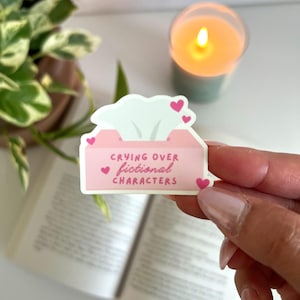 May include: A pink sticker in the shape of a tissue box with the text "Crying over fictional characters" and small pink hearts. The sticker is held in front of an open book, a candle, and a plant.