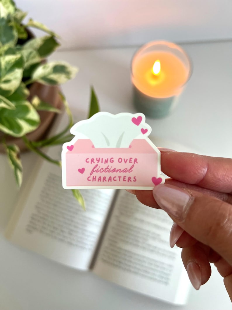 May include: A pink sticker in the shape of a tissue box with the text "Crying over fictional characters" and small pink hearts. The sticker is held in front of an open book, a candle, and a plant.