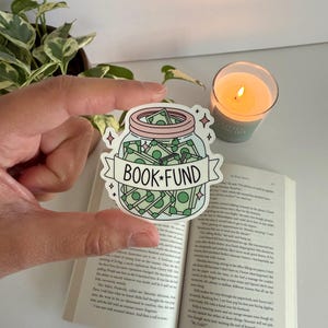 May include: A sticker featuring a jar filled with money, labeled "BOOK FUND" on a banner. The sticker is held over an open book, with a lit candle and a plant in the background. The jar is light blue with a pink lid.