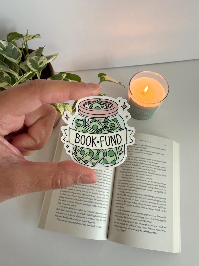 May include: A sticker featuring a jar filled with money, labeled "BOOK FUND" on a banner. The sticker is held over an open book, with a lit candle and a plant in the background. The jar is light blue with a pink lid.