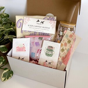 May include: A gift box filled with book-themed items. Includes a canvas tote bag, an iced coffee sleeve, a pink book-patterned pouch, stickers, a glass jar with a straw, and a bookmark with a tassel. The box is white with a brown cardboard lid.