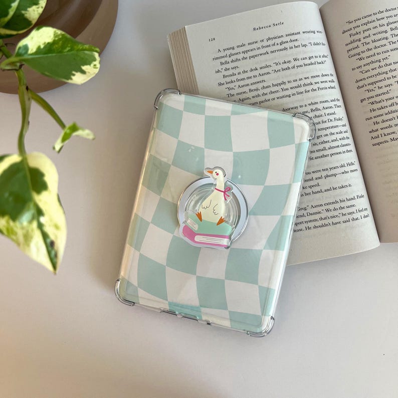 May include: A phone case with a light blue and white checkered pattern. A clear ring holder with a cartoon goose design is attached. The goose is wearing a pink bow and sitting on a book. The case is next to an open book.