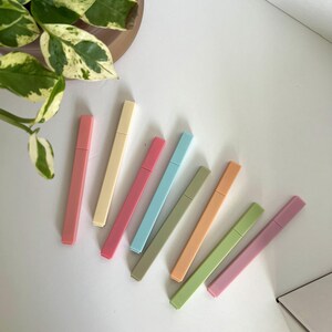 May include: A collection of eight pastel-colored highlighters arranged diagonally on a white surface. The highlighters are in shades of pink, yellow, blue, green, and orange. A potted plant with green and white leaves is in the background.