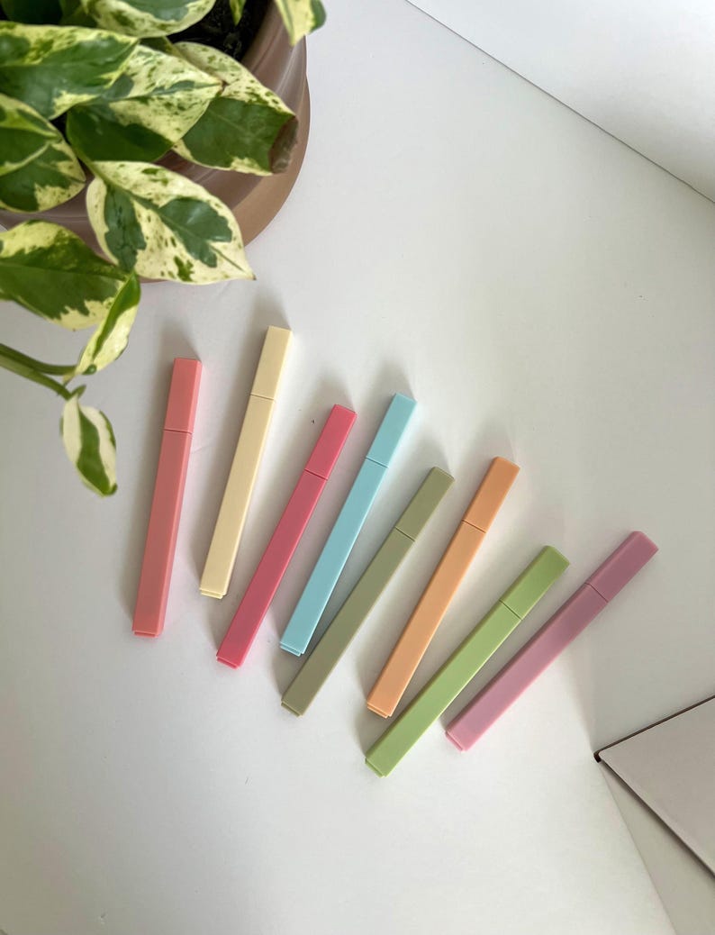 May include: A collection of eight pastel-colored highlighters arranged diagonally on a white surface. The highlighters are in shades of pink, yellow, blue, green, and orange. A potted plant with green and white leaves is in the background.