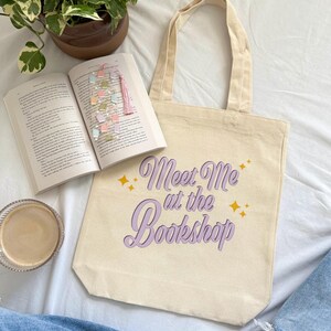 May include: A beige tote bag with the text "Meet Me at the Bookshop" in purple script and gold stars. An open book with a colorful bookmark and a cup of coffee are next to the bag. A potted plant is in the background.