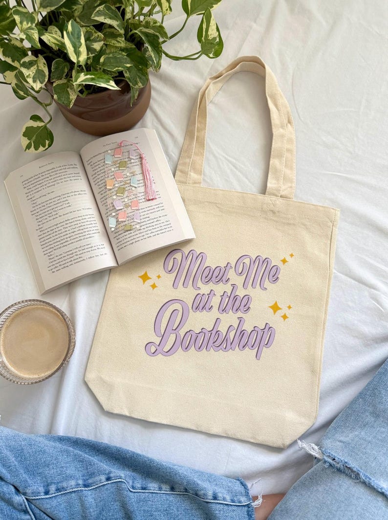 May include: A beige tote bag with the text "Meet Me at the Bookshop" in purple script and gold stars. An open book with a colorful bookmark and a cup of coffee are next to the bag. A potted plant is in the background.