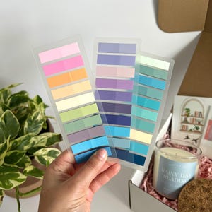 May include: A hand holds three clear plastic sheets of colorful sticky notes in shades of pink, orange, green, blue, and purple. A gift box with a candle, coaster, and card is in the background. The candle has the text "Rainy Day Reading".