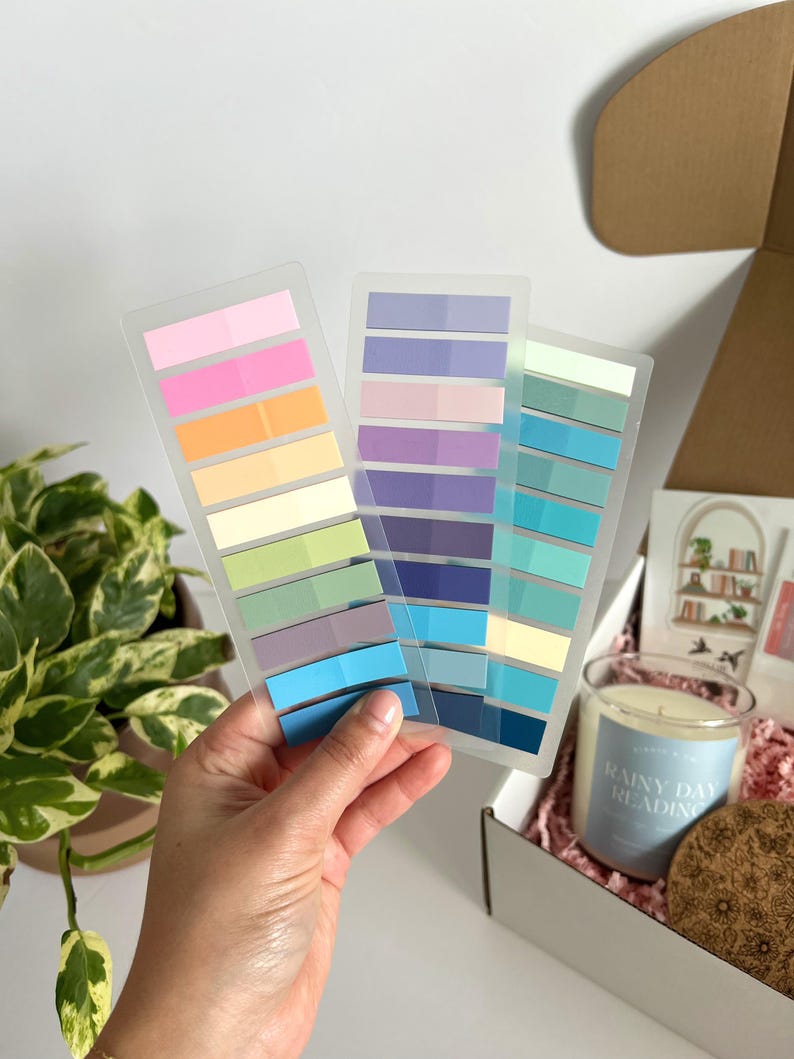May include: A hand holds three clear plastic sheets of colorful sticky notes in shades of pink, orange, green, blue, and purple. A gift box with a candle, coaster, and card is in the background. The candle has the text "Rainy Day Reading".