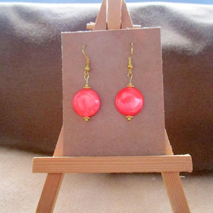 Peut inclure: Une paire de boucles d'oreilles rondes de couleur corail avec des crochets dorés et de petits accents dorés. Les boucles d'oreilles sont présentées sur une carte marron, sur un petit chevalet en bois.