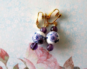 Puede incluir: Pendientes de clip con cuentas de cerámica blanca con diseños florales azules, acentuados por cuentas de amatista púrpura. Los pendientes tienen clips dorados. El fondo tiene un diseño floral.