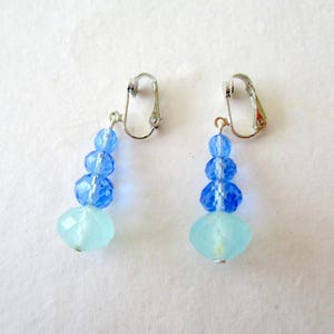 Op de afbeelding: Een paar clip-oorbellen met een cascade van blauwe en lichtblauwe facetkralen. De oorbellen hebben een zilverkleurige clip en een druppelvorm, met een grotere, lichtblauwe kraal aan de onderkant.