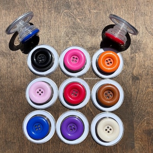 May include: A selection of colourful phone grips with button designs. The grips feature a variety of colours, including black, pink, orange, red, brown, blue, purple, and white. Each grip has a clear, collapsible stand.