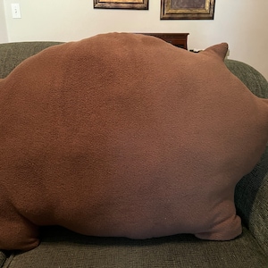 Puede incluir: Un gran cojín de peluche marrón con forma de oso. El cojín está sentado en un sofá verde.