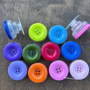 May include: A collection of eleven circular phone grips designed to resemble colorful buttons. The grips feature four central holes and come in various solid colors including lime green, blue, purple, olive green, red, orange, pink, and lavender.