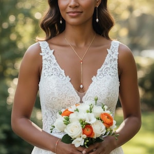 Op de afbeelding: Een bruid in een witte kanten trouwjurk houdt een boeket witte en oranje bloemen vast. Ze draagt een gouden ketting met een parelhanger, bijpassende oorbellen en een armband. De achtergrond is wazig.