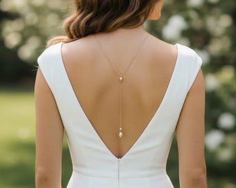 May include: A woman in a white sleeveless dress with a deep V-back. A delicate gold necklace with two pearls hangs down the back. The background is blurred with greenery.
