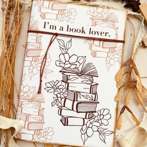 Può includere: Un diario bianco con la scritta "I'm a book lover" e una pila di libri con decorazioni floreali. Il diario è legato con uno spago marrone e circondato da elementi decorativi.