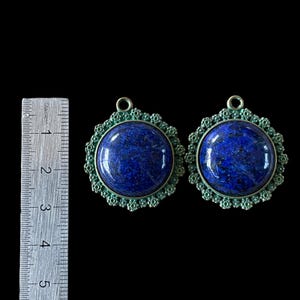 Op de afbeelding: Twee blauwe lapis lazuli stenen hangers met sierlijke groene metalen frames. De hangers zijn ongeveer 3,8 cm in diameter.
