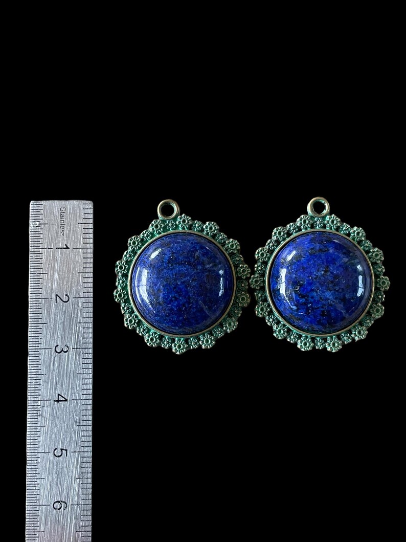 Op de afbeelding: Twee blauwe lapis lazuli stenen hangers met sierlijke groene metalen frames. De hangers zijn ongeveer 3,8 cm in diameter.