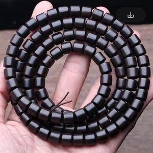 Könnte beinhalten: Eine schwarze Holzperlenkette mit mehreren Perlensträngen. Die Perlen sind zylindrisch geformt und haben eine glatte, polierte Oberfläche.