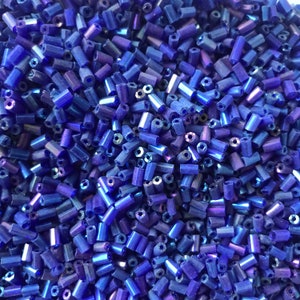 Può includere: Una vista ravvicinata di una grande quantità di piccole perline di vetro blu iridescente. Le perline sono di forma cilindrica e hanno una leggera lucentezza metallica.