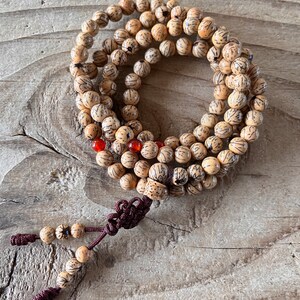 Op de afbeelding: Een beigehouten mala-ketting met een bruine kwast. De kralen zijn rond en hebben een gesneden ontwerp. Er zijn ook een paar rode kralen verspreid over de ketting.