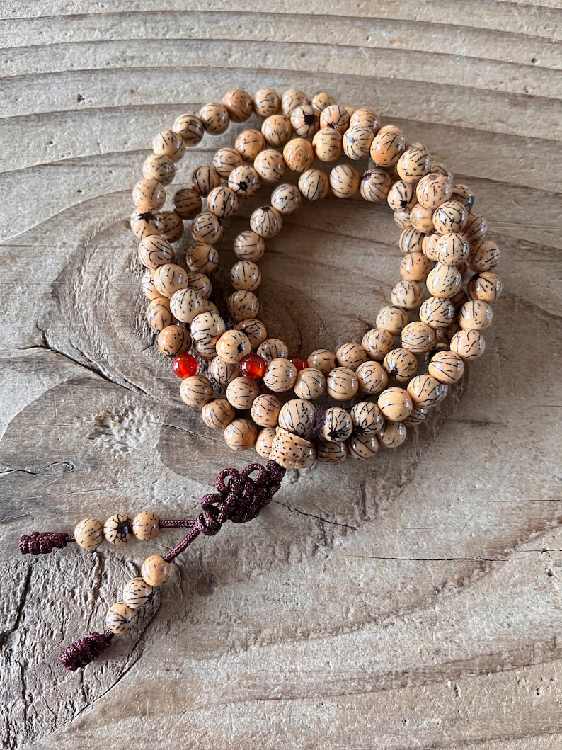 Op de afbeelding: Een beigehouten mala-ketting met een bruine kwast. De kralen zijn rond en hebben een gesneden ontwerp. Er zijn ook een paar rode kralen verspreid over de ketting.