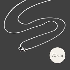 Può includere: Una collana a catena a serpente color argento con chiusura a moschettone. La collana è lunga 70 cm e poggia su uno sfondo grigio scuro. La catena ha una superficie liscia e riflettente.