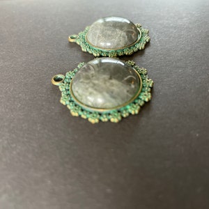 Op de afbeelding: Twee ronde, zilverkleurige metalen bedels met een groene emaille rand. Elke bedel heeft een heldere glazen cabochon met een grijs-wit patroon.