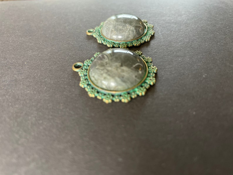 Op de afbeelding: Twee ronde, zilverkleurige metalen bedels met een groene emaille rand. Elke bedel heeft een heldere glazen cabochon met een grijs-wit patroon.
