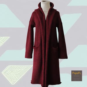 May include: A long, burgundy hooded coat with two front pockets. The coat is made of a soft, textured material and is worn over a white roll neck. The coat has a relaxed fit and a long length, suitable for cooler weather.