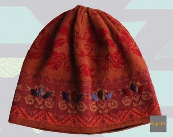 May include: A knitted beanie in shades of orange and red, featuring a floral pattern. The hat has a decorative border with stylized floral and leaf motifs in purple, blue, and orange. The beanie is a winter accessory.