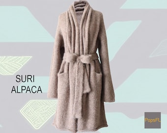 May include: A long-sleeved, knee-length beige cardigan with a shawl collar, two front pockets, and a matching tie belt at the waist. The text "SURI ALPACA" is printed on the left, and the text "PopsFL" is in a gray box in the bottom right corner.