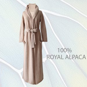 May include: A long, beige knit robe featuring a shawl collar, a matching tie belt at the waist, and two front pockets. The garment has a ribbed turtleneck collar underneath and includes the text 100% ROYAL ALPACA printed on the right side.