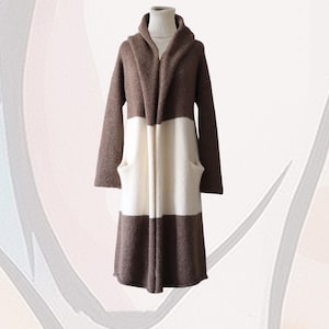 May include: A long-sleeved, knee-length cardigan featuring a colour-blocked design with horizontal bands of brown and cream. The garment has a wide shawl collar, an open front, and two side pockets, worn over a cream-coloured turtleneck jumper.