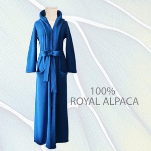 May include: A floor-length, royal blue hooded robe featuring long sleeves, two front pockets, and a matching tie belt at the waist. The garment is worn over a beige ribbed turtleneck sweater. The text 100% ROYAL ALPACA is visible on the right.