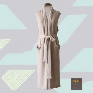 May include: A long, sleeveless beige cardigan with a shawl collar and a matching belt tied at the waist. The cardigan has two front pockets and is displayed on a mannequin. The background has a light grey colour with abstract shapes.