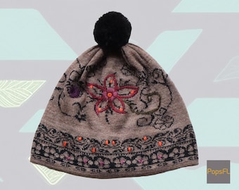 May include: A brown knitted beanie with a black pom pom and embroidered floral design. The beanie has a black and orange trim.