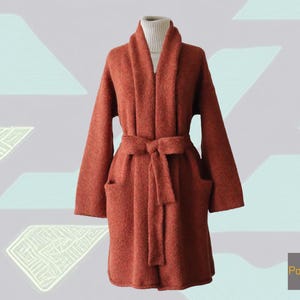 May include: A rust-coloured, long-sleeved robe with a shawl collar, a waist tie, and two front pockets. The robe is displayed on a mannequin and has a white turtleneck underneath. The robe is made of a soft, textured material.