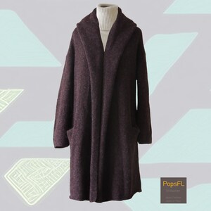 Könnte beinhalten: Ein langer, dunkelburgunderfarbener Mantel mit Kapuze und zwei Vordertaschen. Der Mantel besteht aus einem dicken, strukturierten Material und wird über einem weißen Rollkragenpullover getragen. Der Mantel hat eine lockere Passform und einen Schalkragen.