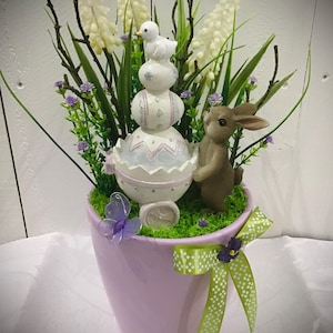 May include: A purple ceramic planter with a green and white ribbon bow. The planter is filled with green moss, white flowers, and a small white bunny figurine. The bunny is holding a stack of pastel Easter eggs.