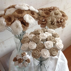 Può includere: Tre bouquet di fiori di juta e pizzo di diverse dimensioni. I bouquet sono disposti in barattoli di vetro su una tovaglia bianca. I fiori sono un mix di marrone e bianco.